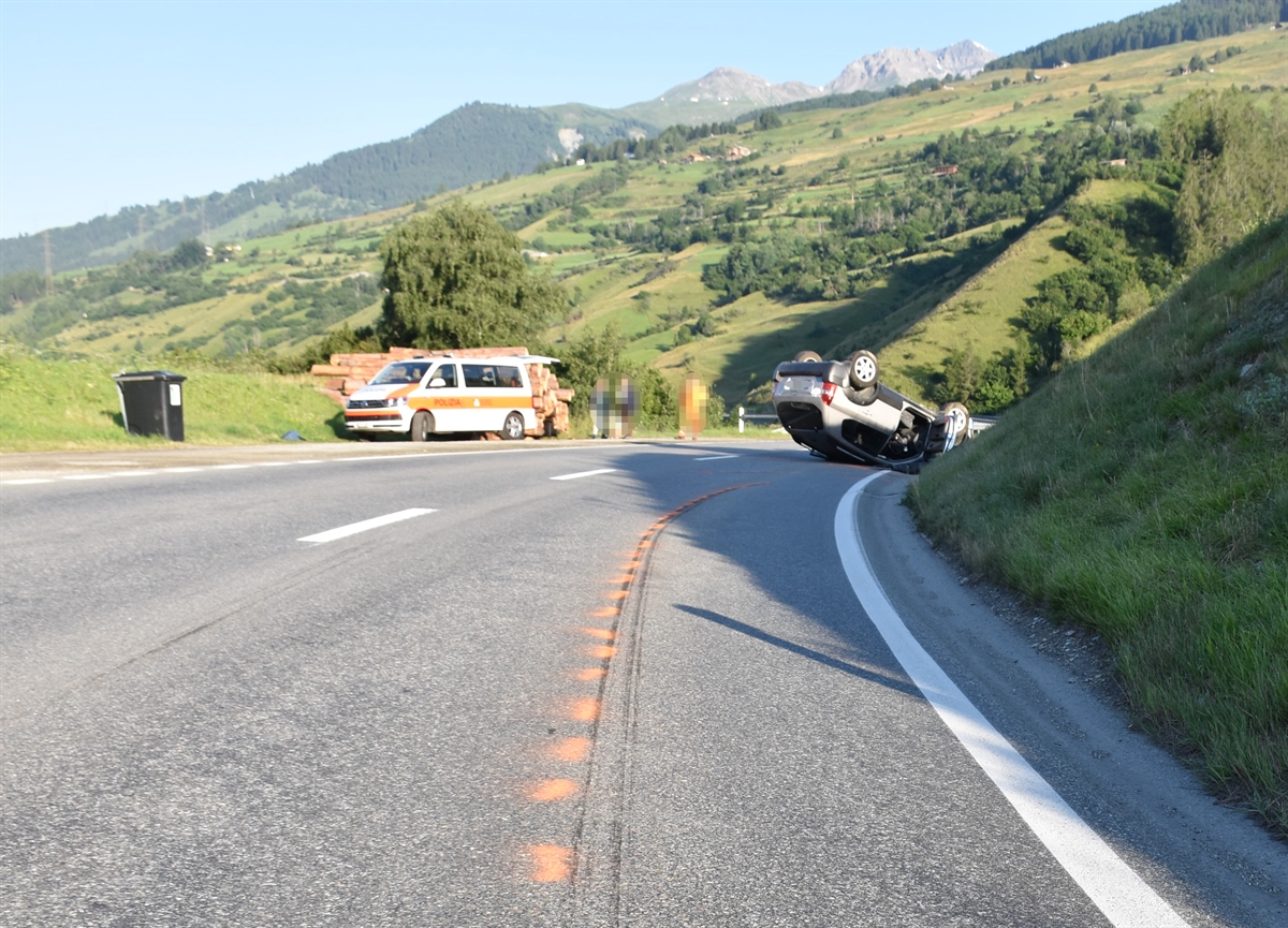 Rechtskurve von Crusch in Richtung Scuol. Im Hintergrund Berge und blauer Himmel. Auf der linken Seite ein Polizeiauto und rechts das auf dem Dach liegende Auto.