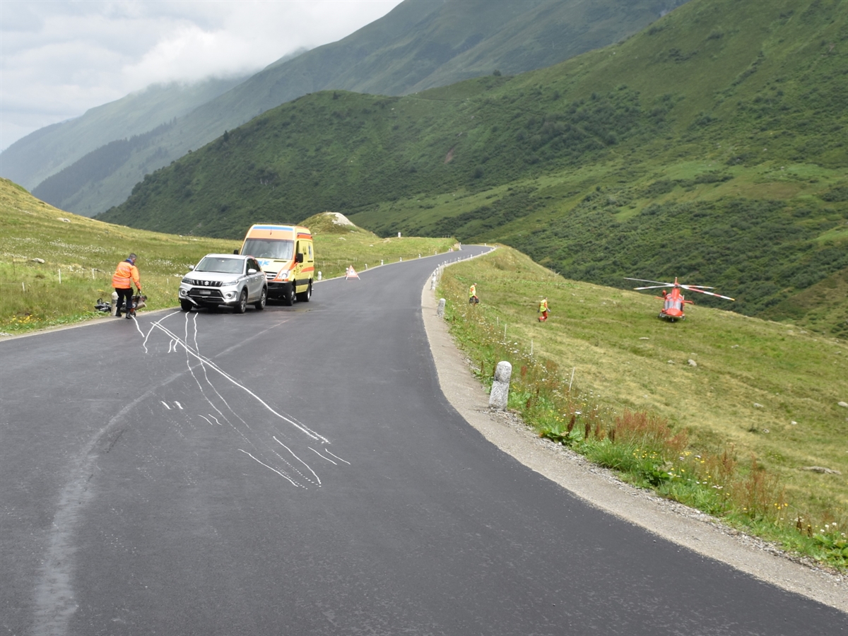 Rechtskurve der Oberalpstrasse. Auf der Gegenfahrbahn liegt das Motorrad neben der Strasse. Ein Polizist zeichnet die Spuren auf der Fahrbahn ein. Dahinter steht der Personenwagen sowie hinter diesem das Ambulanzfahrzeug. Auf der rechten Bildseite steht in der Wiese der Helikopter der Rega.