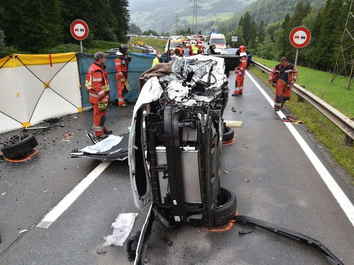 Das auf der Seite liegende Auto. Links stehen zwei Feuerwehrleute und schirmen mit einem Sichtschutz den Einblick auf das Fahrzeug ab. Im Hintergrund stehen