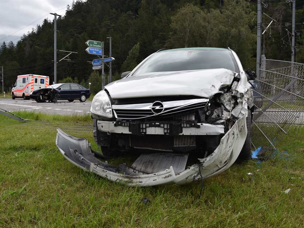 Frontaufnahme des einen Autos. Es ist frontal und seitlich links stark beschädigt. Dasselbe mit dem links davon im Hintergrund stehenden anderen unfallbeteiligten Autos. Links davon ein Ambulanzfahrzeug. 