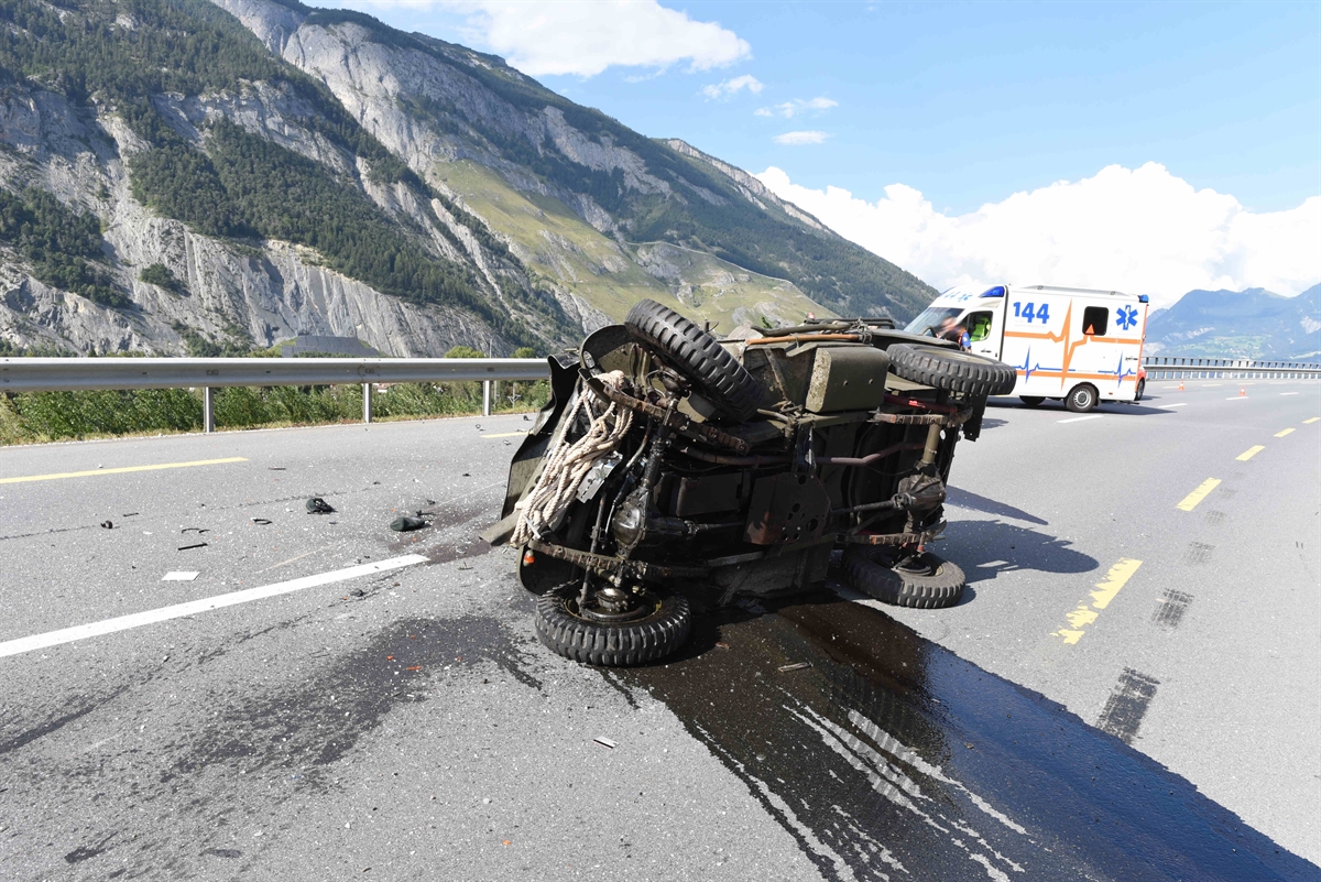 Ein älterer Willys USA Personenwagen liegt auf der Seite auf der Strasse und im Hintergrund die Ambulanz.