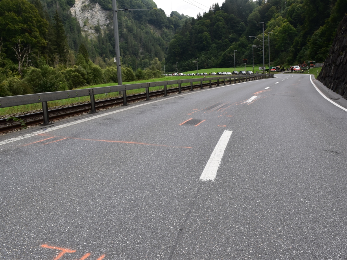 Auf der Strasse mit oranger Farbe eingezeichnete Unfallspuren. Rechts eine Stützmauer, linke eine Leitplanke und dahinter das Geleise der Rhätischen Bahn. Im Hintergrund zwei Polizeiautos und eine Autokolonne.