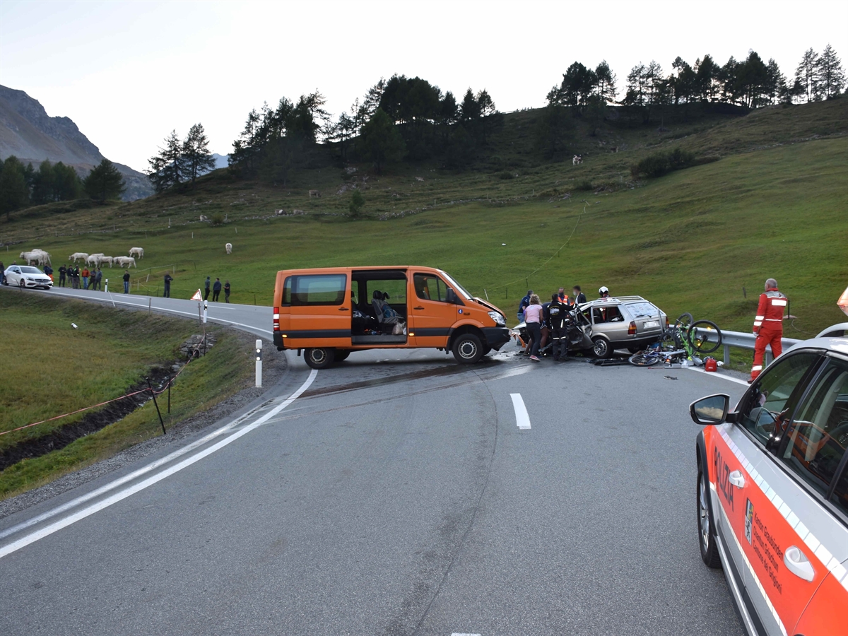 Rechts ein Polizeiauto. In Bildmitte die Kurve bergwärts gesehen nach links. In der Kurve rechts der stark deformierte Personenwagen, links davon der Kleinbus quer auf der Strasse. Rettungskräfte und weitere Helfer im Einsatz.