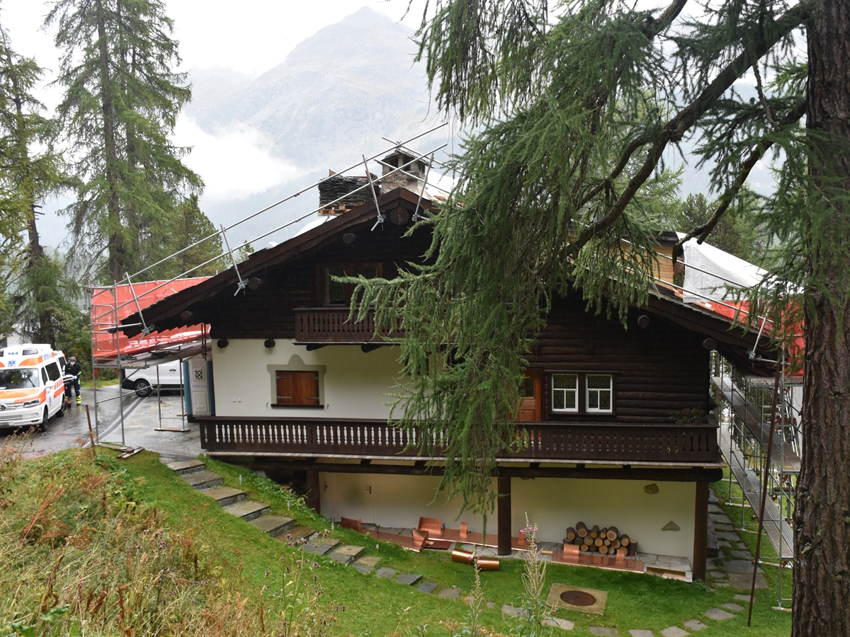 Ein von Bäumen umsäumtes Haus. Im Hintergrund Berge. Links vom Haus ein Ambulanzauto. Auf dem Dach des Hauses sowie der Fassade links und rechts Metallgerüste.