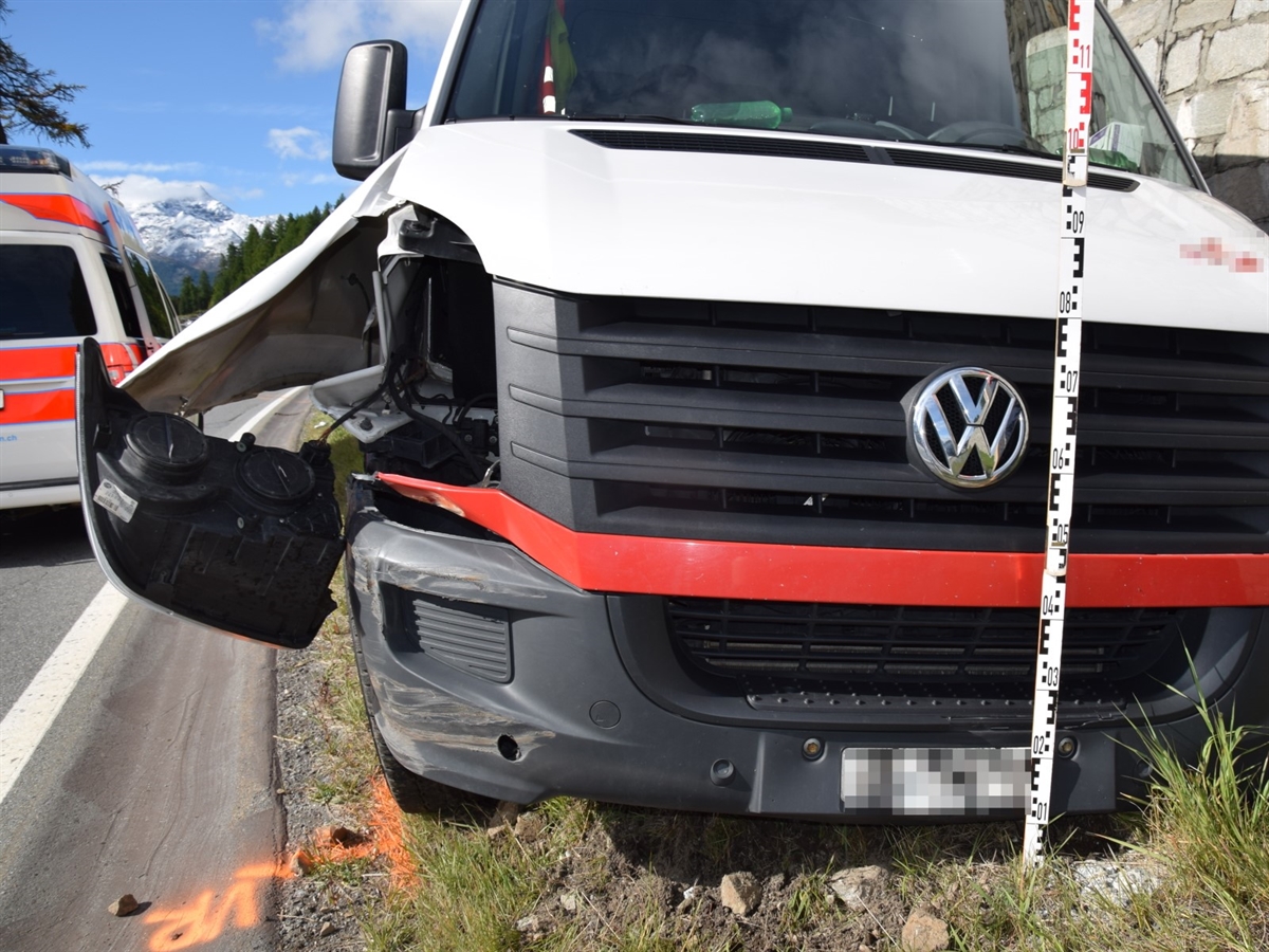 Der Lieferwagen von vorne. Er steht an der Stützmauer und weist eine beschädigte Front auf. Links davon ein Ambulanzfahrzeug. Im Hintergrund Berge und blauer Himmel. 