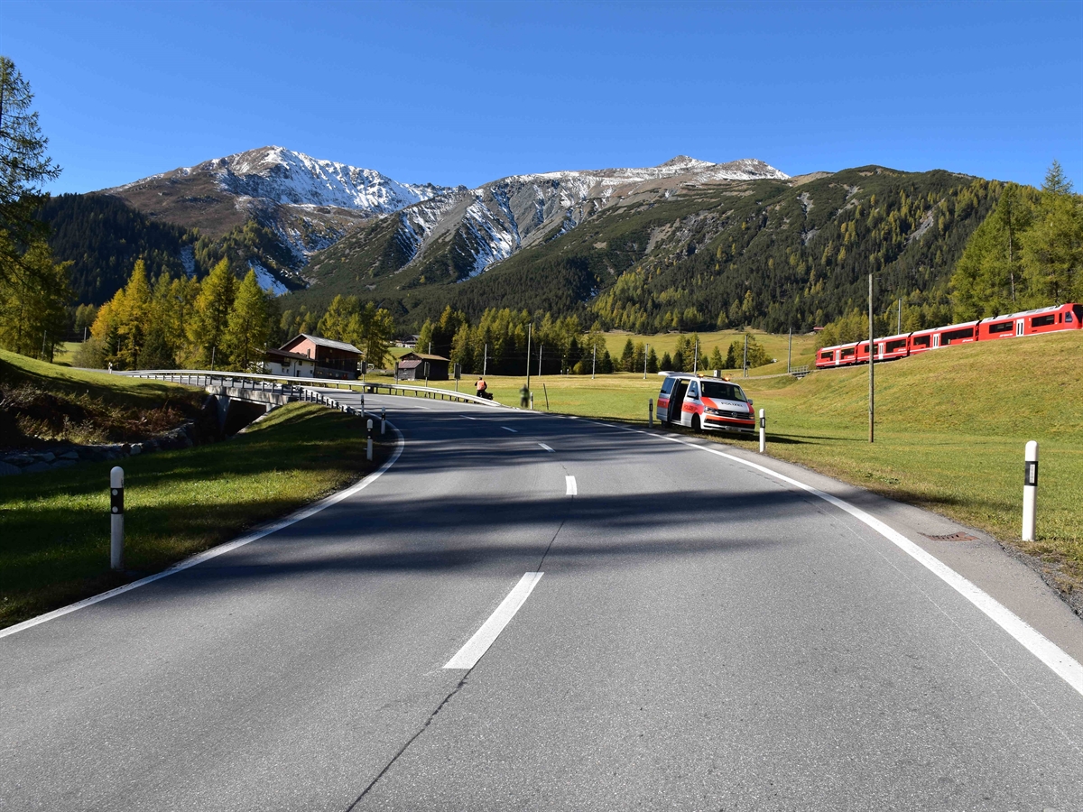 Die Linkskurve. In Bildmitte ein Polizist an der Strasse, rechts davon ein Polizeiauto. Weiter rechts ein wenig entfernt ein Zug der Rhätischen Bahn RhB. Im Hintergrund Berge und blauer Himmel.