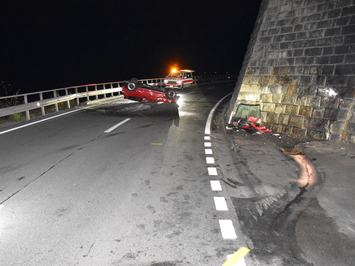 Die Unfallstelle mit der rechtsseitigen Stützmauer. Vor dieser liegen Fahrzeugtrümmer. Links davon liegt das Auto auf dem Dach und hinter diesem ein Patrouillenfahrzeug der Kapo GR mit eingeschaltetem gelben Blinklicht.