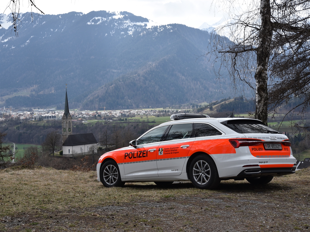 Ein Polizeiauto auf einem Kiesplatz. Im Hintergrund eine Kirche und weiter entfernt ein Dorf. 