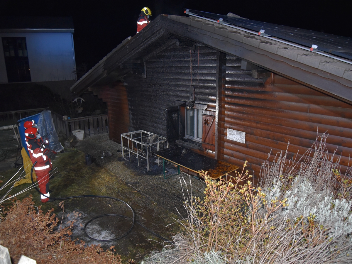 Links auf einer Grünfläche stehen zwei Feuerwehrleute in Bereitschaftsstellung mit Wasserschläuchen. Rechts davon das Haus mit stark angesengter Holzfassade. Oben auf dem Giebeldach ein Feuerwehrmann.
