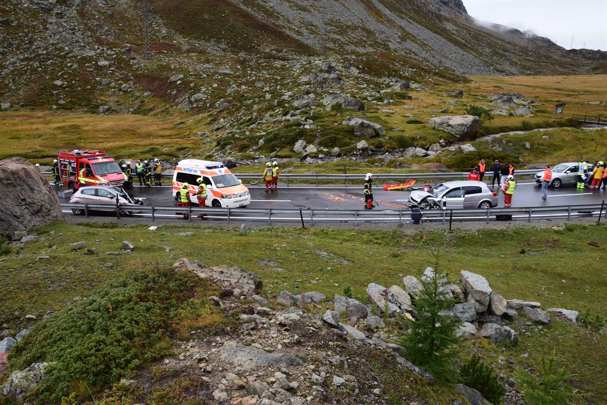 Strassenverlauf der Nationalstrasse N29 im Bereich der Unfallstelle, Höhe Alp Güglia. Auf der linken Seite ist ein Einsatzfahrzeug der Feuerwehr und der Sanität sowie ein beteiligtes Unfallfahrzeug zu erkennen. Auf der rechten Seite ist das zweite Unfallfahrzeug zu erkennen. Zudem sind diverse Rettungskräfte von Polizei, Feuerwehr und Sanität zu erkennen.