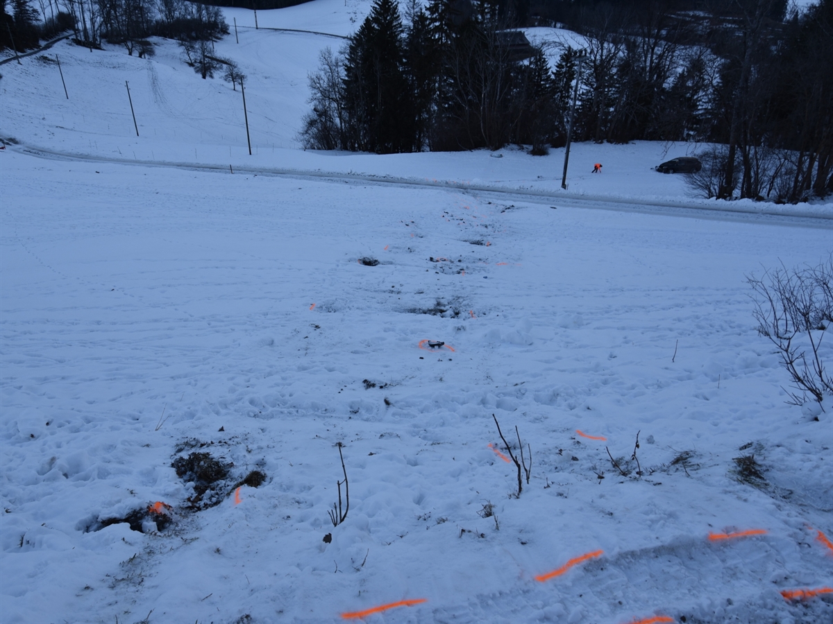 Der verschneite Abhang mit den Überschlagsspuren im Schnee. In der Ferne markiert ein Polizist die Spuren. Neben einer Baumgruppe steht das Unfallfahrzeug.