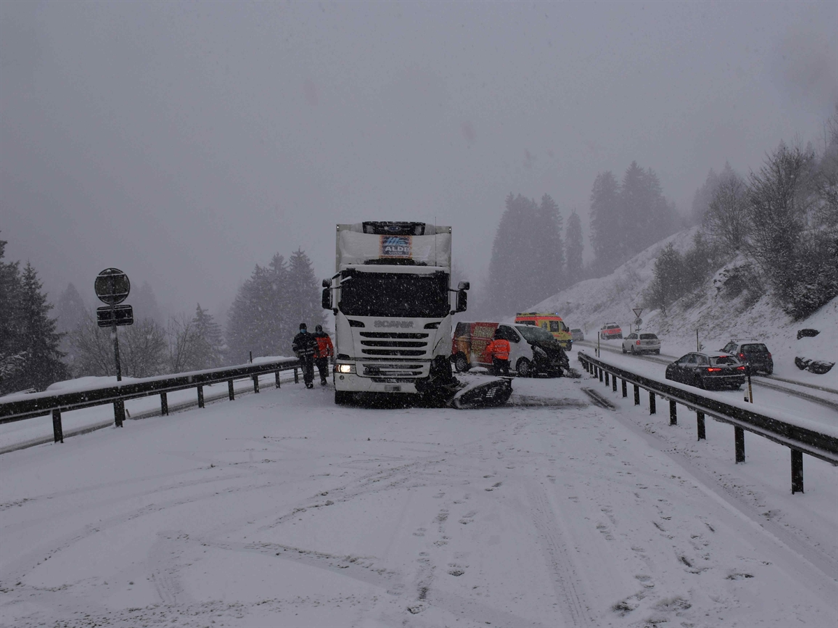 Die verschneite Strasse. Frontal gesehen steht links ein an der linken Front beschädigter Lastwagen, rechts davon im Hintergrund der stark beschädigte Lieferwagen. Hinter dem Lieferwagen ein Ambulanzauto.