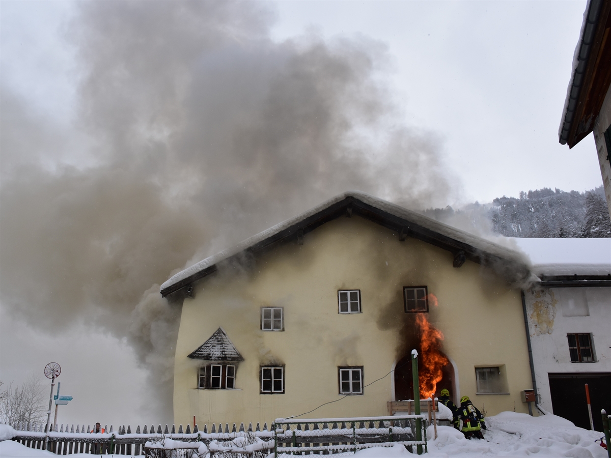 Frontaler Blick auf die Eingangsfassade. Aus dem Eingang steigen Flammen empor, aus dem ganzen Haus quillt Rauch. Vor dem Eingang zwei Feuerwehrleute im Einsatz.