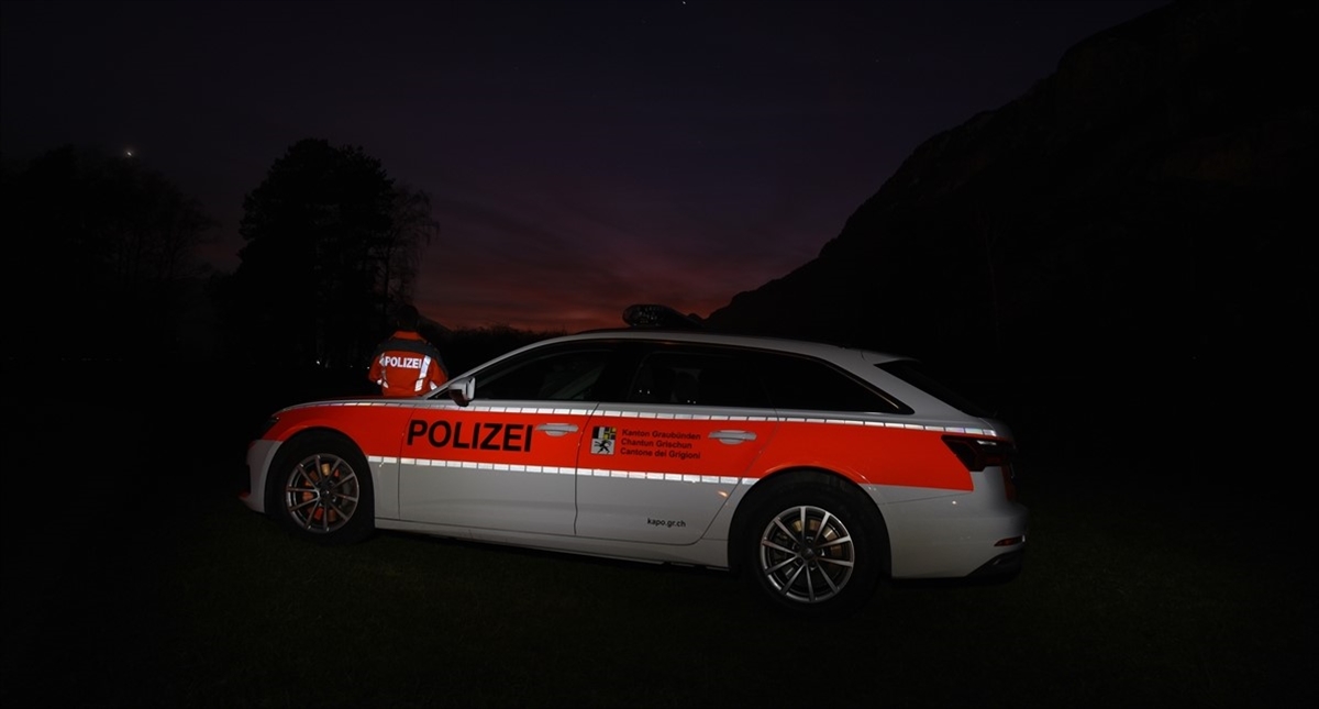 Nachtaufnahme mit Blick auf die linke Seite eines Polizeiautos. Links hinter dem Auto ein Polizist. In der Ferne Berge in der Dämmerung.