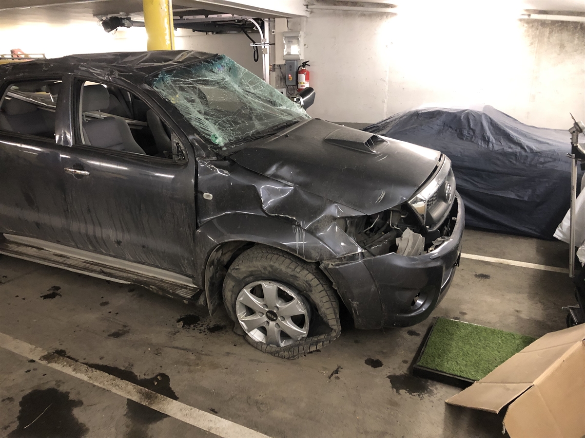 Aufnahme von vorne rechts auf das total beschädigte Fahrzeug. Es steht in einer Garage. Gut erkennbar die eingedrückte Windschutzscheibe, der kaputte Reifen vorne rechts und das eingedrückte Dach.