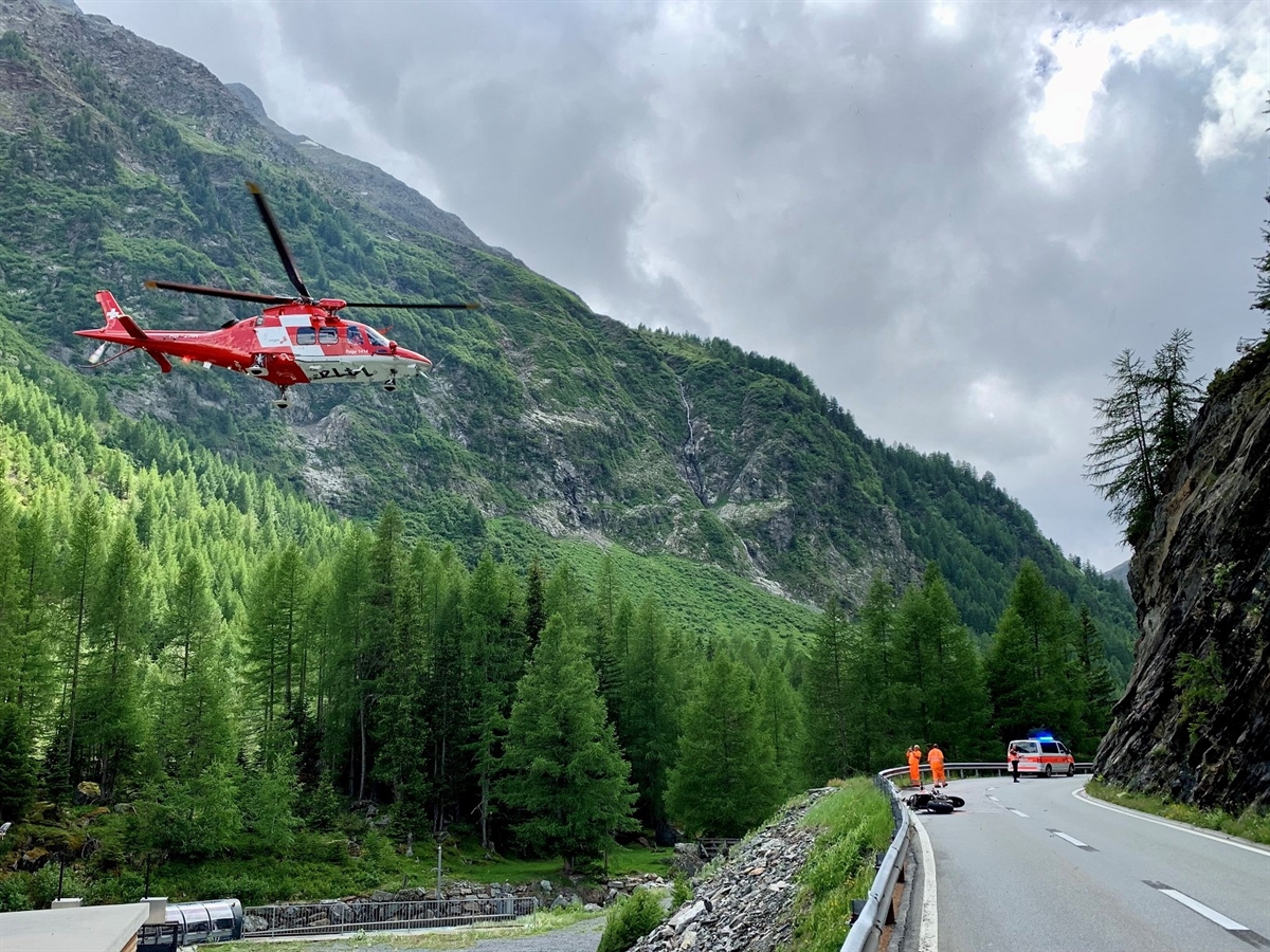Rechts die Unfallstelle, links an der Strasse liegt das Motorrad. Einsatzkräfte und ein Polizeiauto auf der Strasse. Links im Bild der Rega-Helikopter im Flug. Berge grenzen den bewölkten Horizont ab.