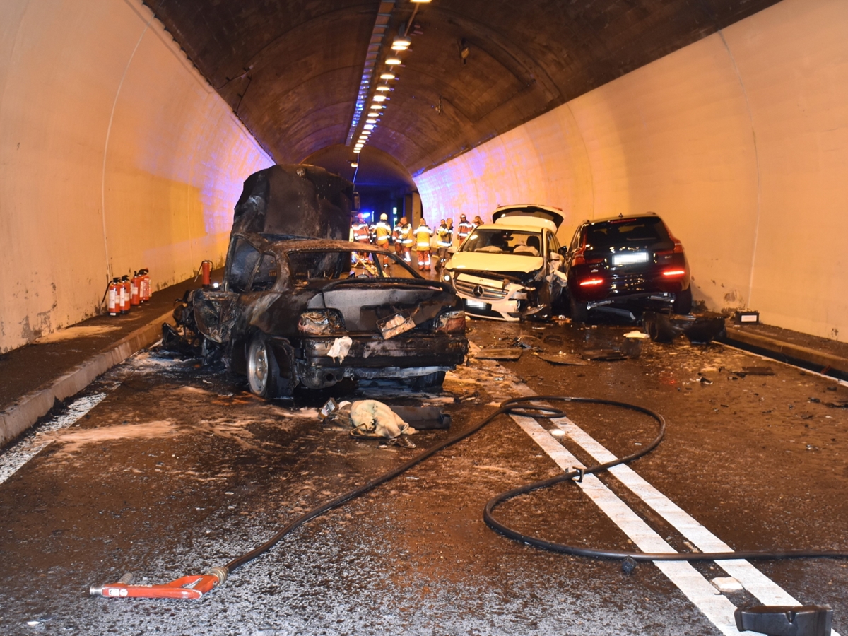 Die Unfallstelle im Tunnel Brusei. Auf der linken Fahrbahn steht das ausgebrannte Fahrzeug. Auf der rechten Fahrbahn die beiden anderen Unfallfahrzeuge. Um die Fahrzeuge herum liegt Feuerlöschmaterial. Im Hintergrund Feuerwehrleute.