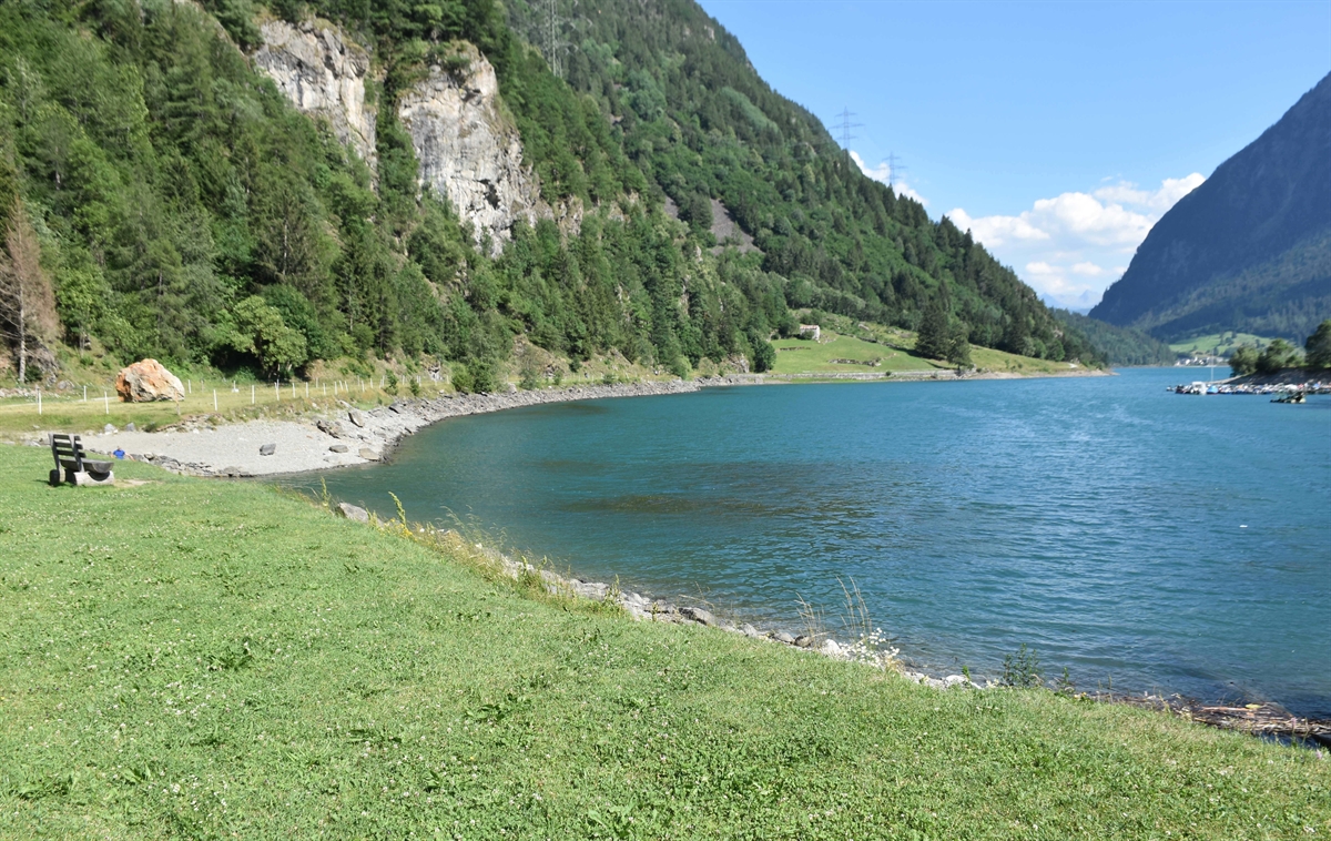 Rechts der See. Im Hintergrund Berge. Links am Seeufer Wiese. 