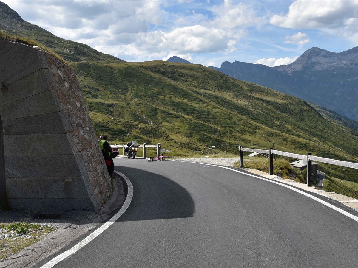 Links eine Galerie und rechts davon die Strasse. An deren rechten Strassenrand liegt ein rosafarbenes Rennrad. Im Hintergrund Berge. 