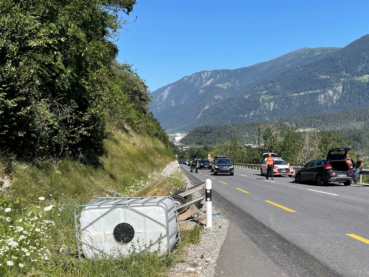 Links neben der Fahrbahn liegen der total zerstörte Anhänger sowie ein Kunststofftank. Auf der talwärts führenden Fahrbahn steht das beschädigte Auto und vor diesem Ein Patrouillenfahrzeug der Kantonspolizei Graubünden. Der blaue Bündnerhimmel rundet das Bild ab.