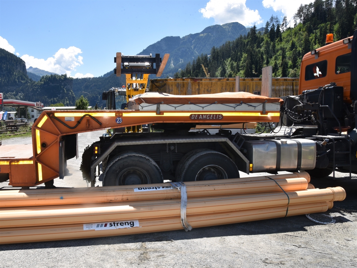Vor dem Satteltransportanhänger liegt ein Rohrpaket. Hinter dem Anhänger steht der Pneulader mit gehobener Gabel.