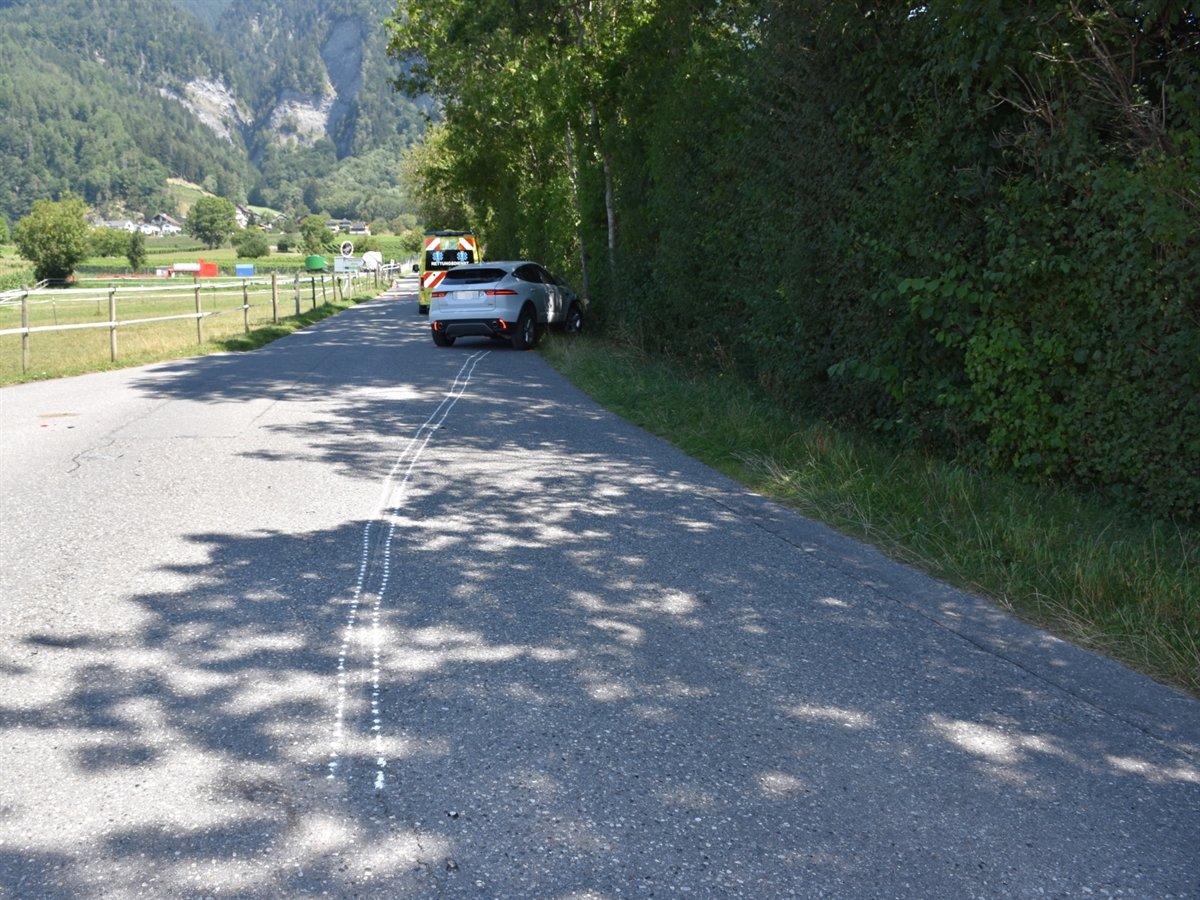 Mitten im Grünen die Verbindungsstrasse nach der Rechtskurve. Angezeichnete Spuren in der Mitte der Strasse. Am Ende der Spur, das Auto der Frau und hinter diesem ein Ambulanzfahrzeug.