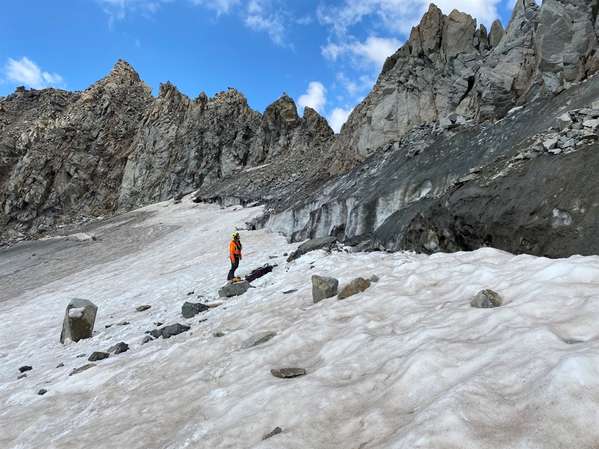 Abfallendes Gelände seitlich aufgenommen. Von Links unten nach rechts oben steigt eine Schneefläche an, geht in einen Bergschrund über und rechts oberhalb Felswände. Im Hintergrund blauer Himmel. Auf der Schneefläche: Eine Einsatzkraft und Steinblöcke.
