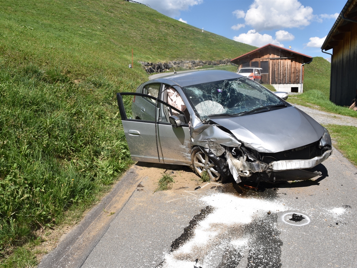 Das total beschädigte Auto steht auf der Strasse zum Hof. Im Hintergrund vor einem Stall steht ein Patrouillenfahrzeug der Kantonspolizei Graubünden. Abgerundete wird das ländliche Bild durch einen wunderbaren blauen Himmel.