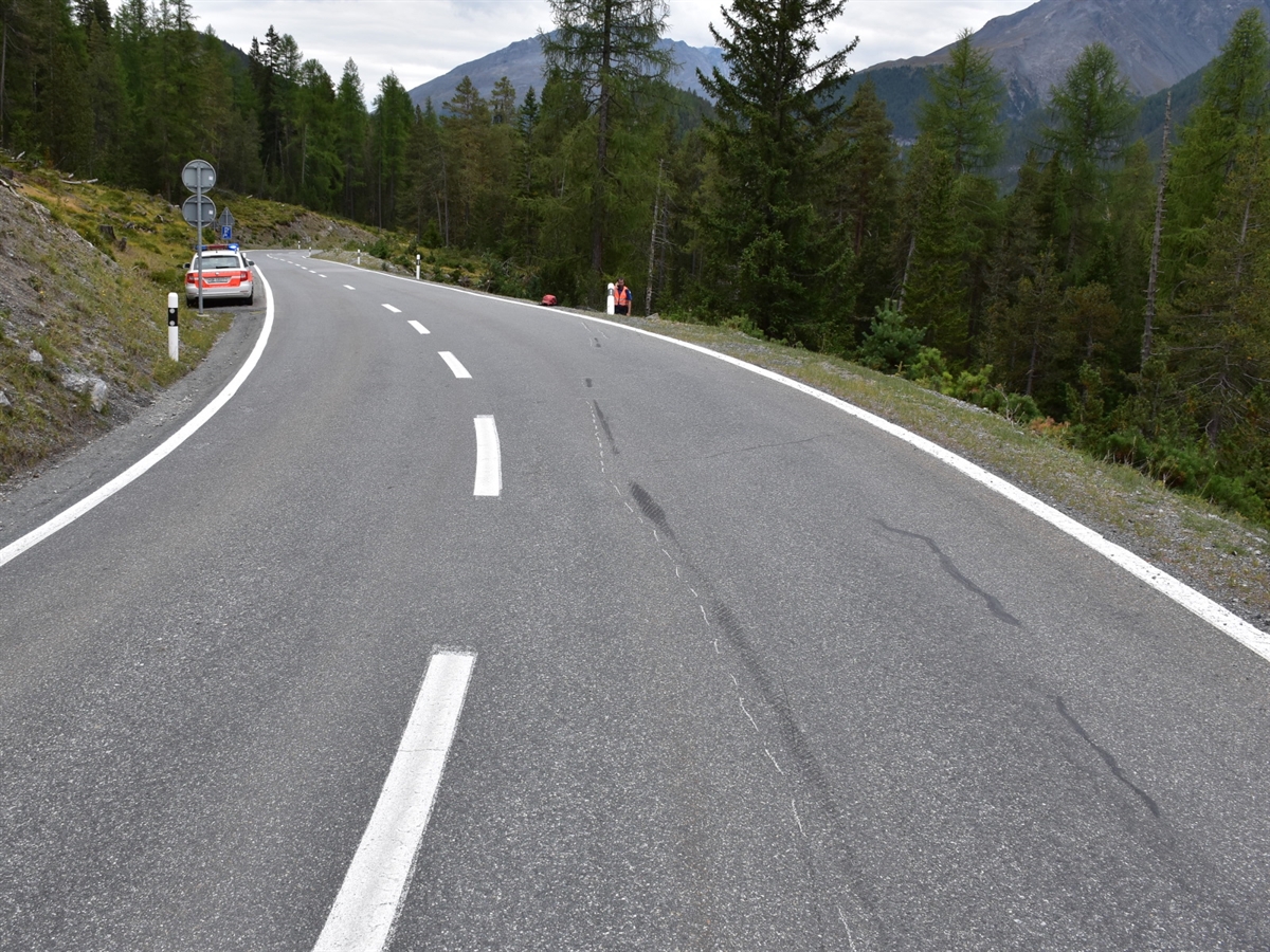 Die von Bäumen umsäumte Linkskurve, in welcher der Motorradfahrer stürzte. Links der Strasse ein Polizeiauto und rechts – in der Flucht der Sturzspuren - ein von einer Böschung auf die Strasse hochlaufender Polizist. 