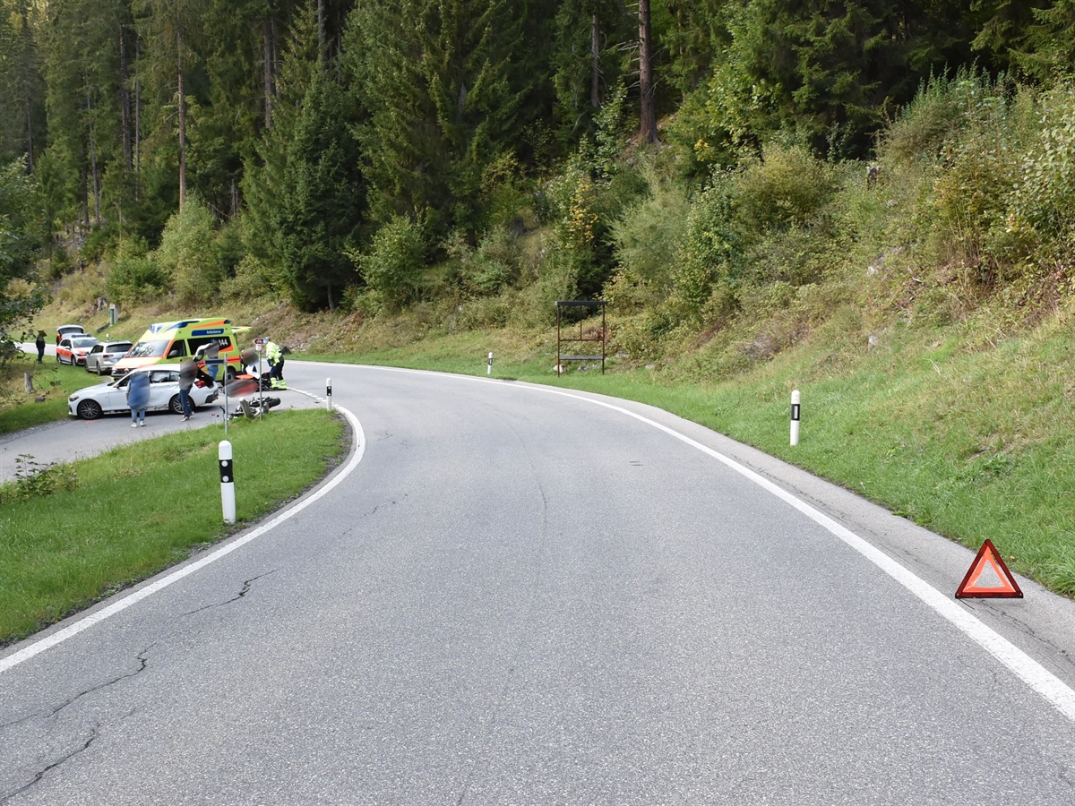 Eine Linkskurve mit einer links abzweigenden Strasse. Auf dieser steht das Unfallauto, liegt das Motorrad und ein Ambulanzfahrzeug.