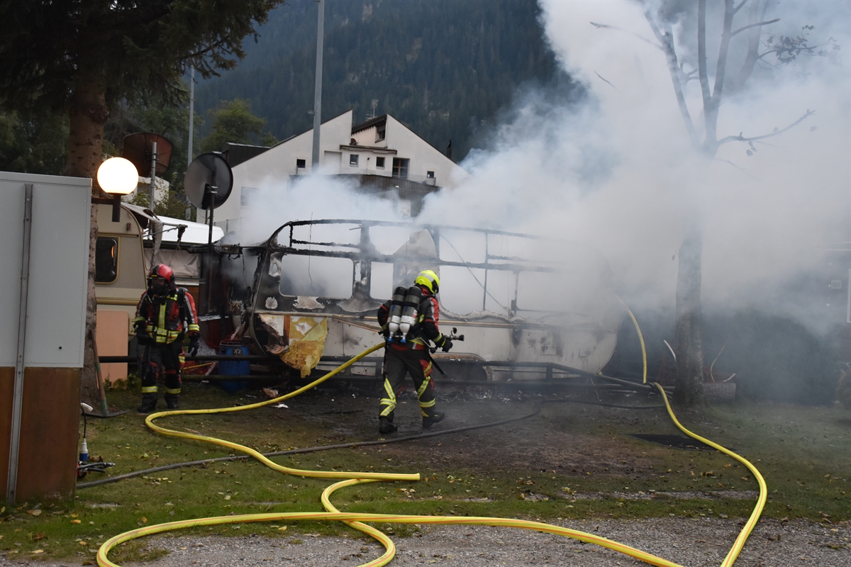 Auf dem Bild ist viel Rauch zu sehen. Der Wohnwagen ist komplett ausgebrannt. Es sind zwei Feuerwehrmänner auf dem Bild. Einer hält noch den Schlauch in der Hand.