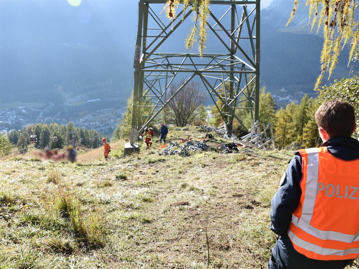 Der Sockelbereich des Hochspannungsmasten. Unter dem Masten Einsatzkräfte die den Verletzten bergen. Im Vordergrund ein Polizist, der den Rettungsvorgang überwacht.