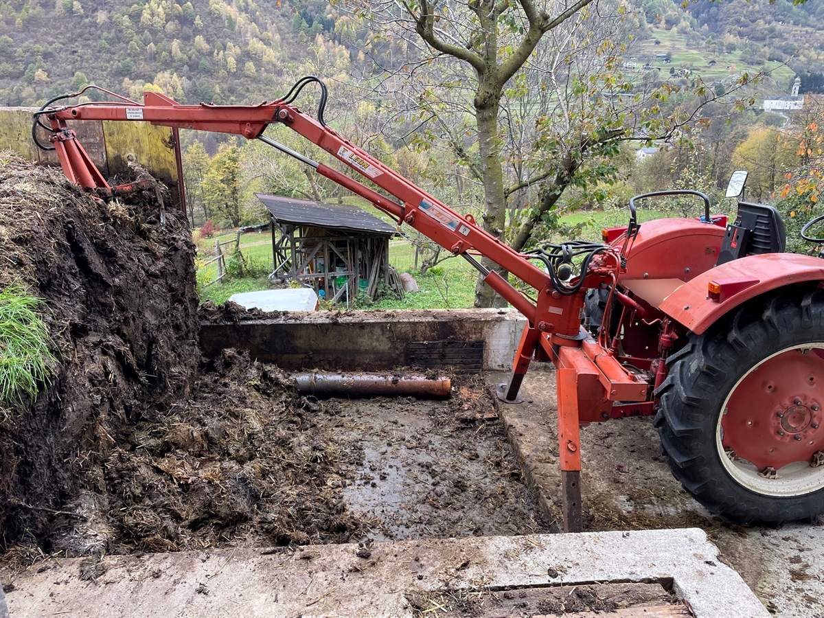 Rechts ein roter Traktor, links daran befestigt der Kran und weiter links der Miststock. Im Hintergrund der Blick ins Tal. 