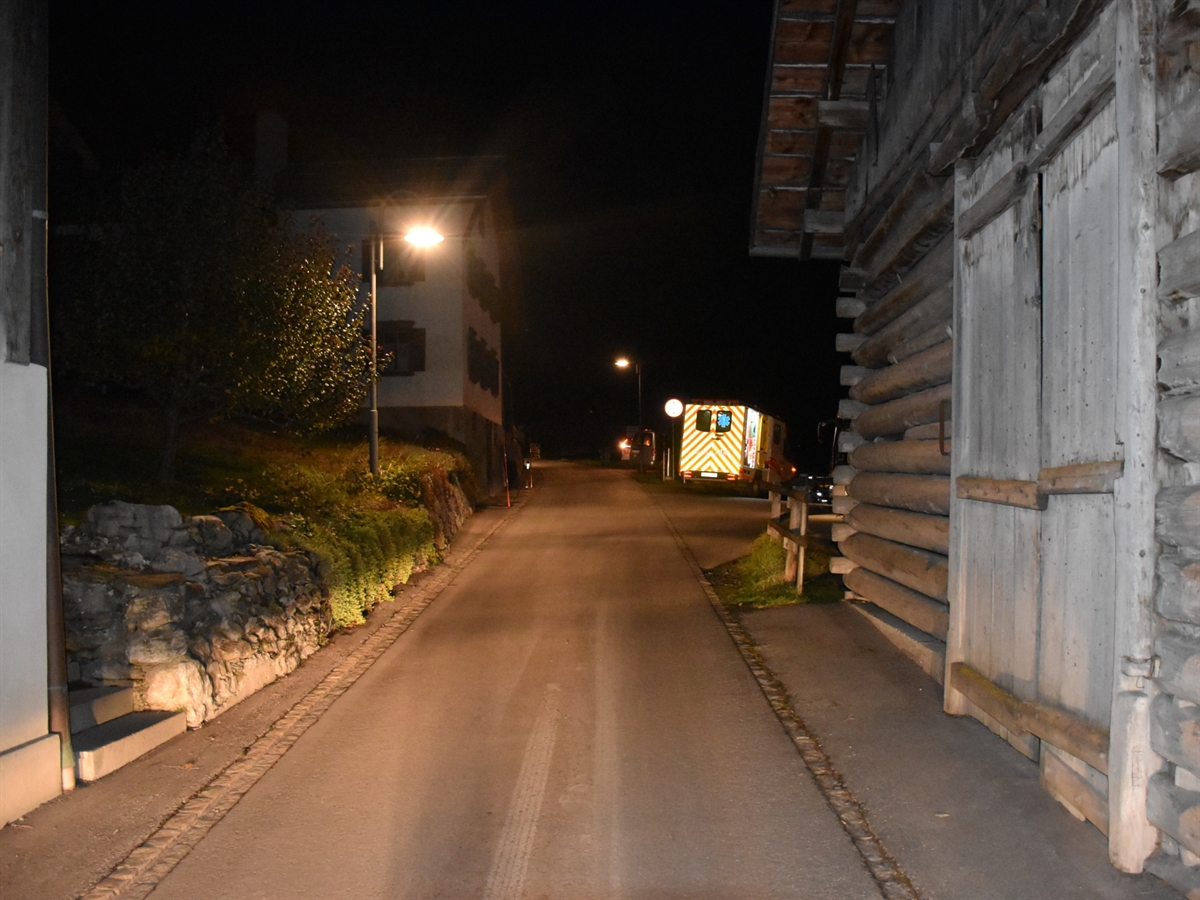 Schmale Strasse in Fidaz. Links und rechts wird die Fahrbahn durch Gebäude eingeengt. Hinter dem Stall auf der rechten Bildseite befindet sich der Wendeplatz. Auf diesem steht ein Ambulanzfahrzeug. Weiter ragt leicht die Front des Linienbusses hinter dem Stall hervor.