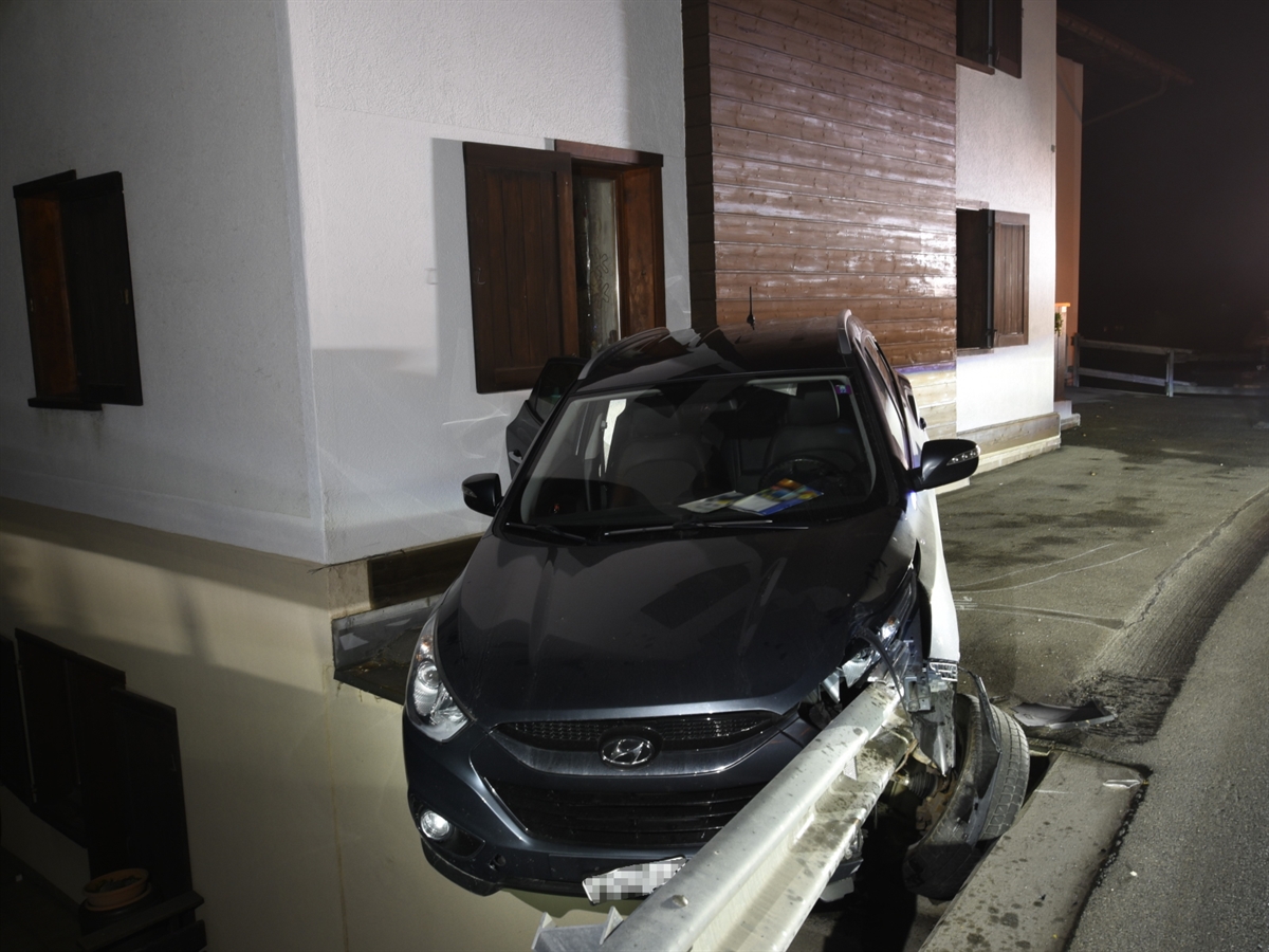 Das beschädigte Auto von vorne. Die Front des Wagens ragt über einen Absatz, welcher um ein Stockwerk nach unten führt. Im Vordergrund die Leitplanke, im Hintergrund ein Haus.