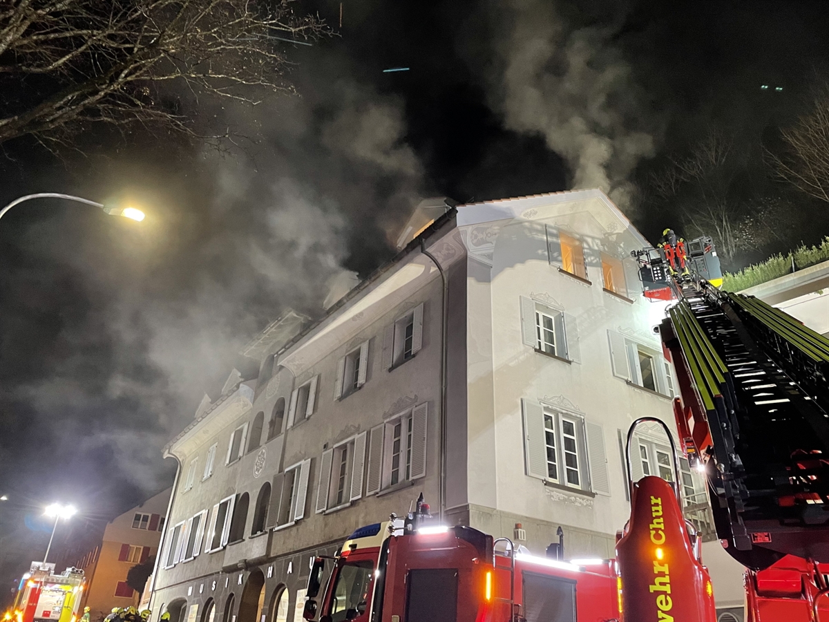 Ein Feuerwehrmann auf einer Autodrehleiter vor einem Fenster des vierstöckigen Mehrfamilienhauses. Rauch steigt in den Nachthimmel.