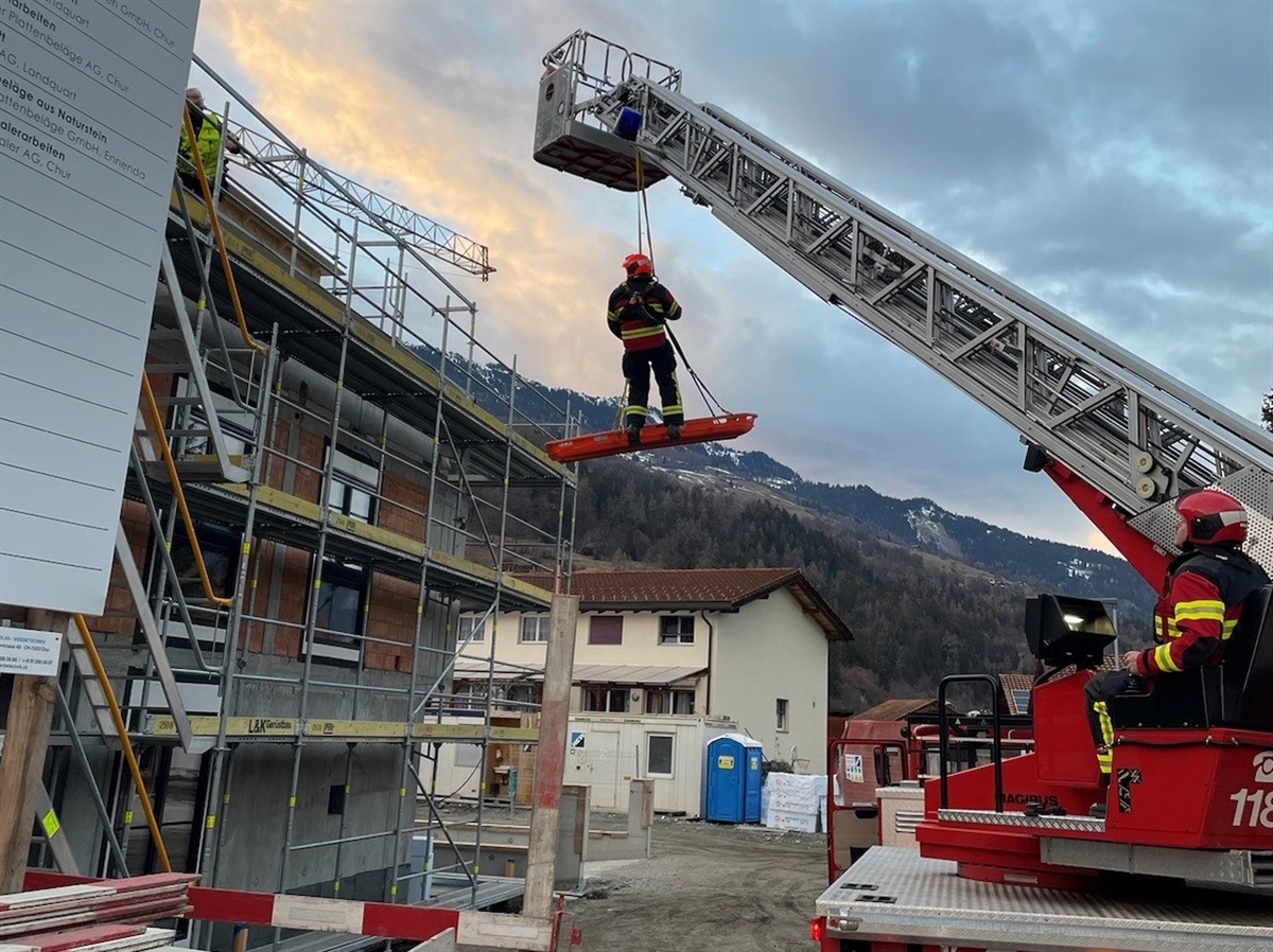 Rechts das Fahrzeug mit dem Bediener der Drehleiter. Diese ist nach links oben zum Neubau hin ausgefahren. Angehängt eine Rettungsbahre, auf der ein Feuerwehrmann steht. 