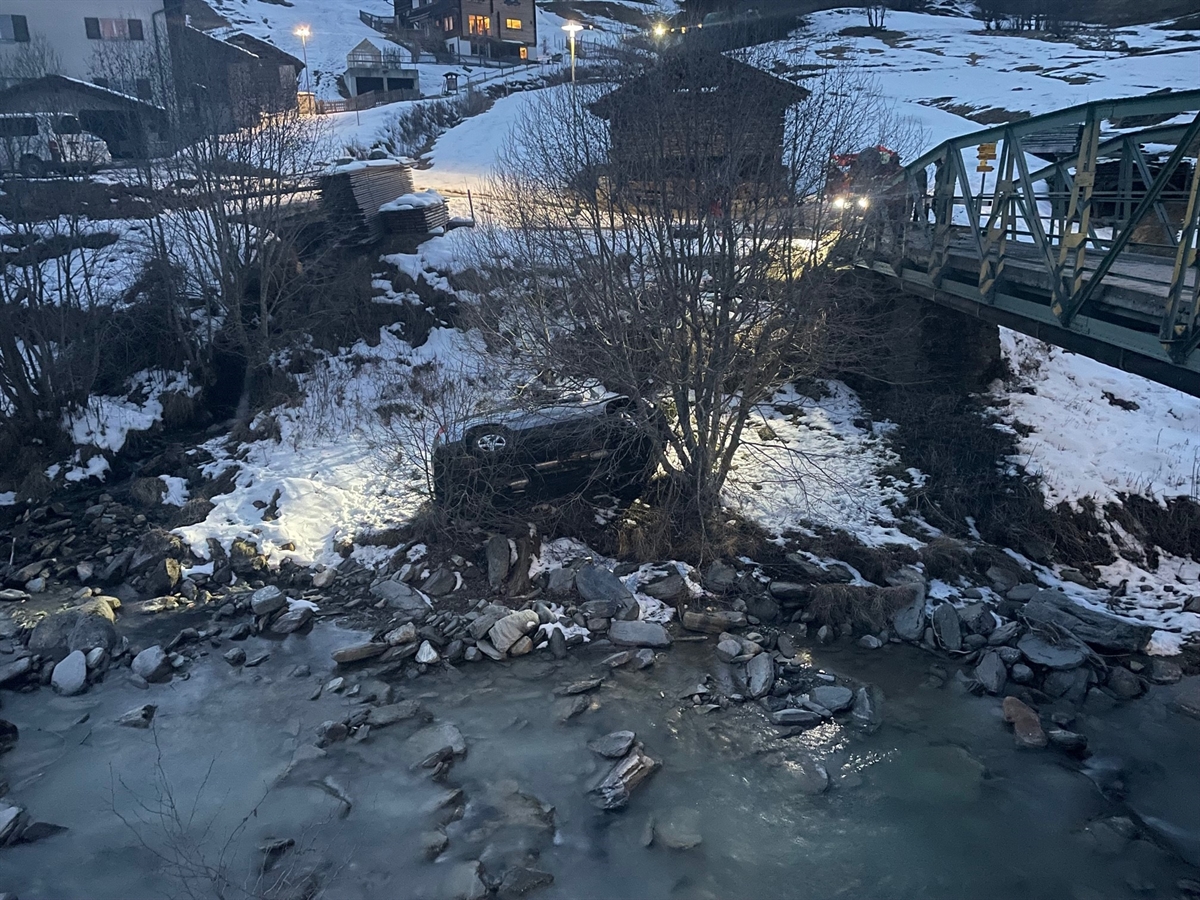 Auto liegt auf der Seite am Uferrand auf Steinen und Schnee, im Hintergrund Häuser