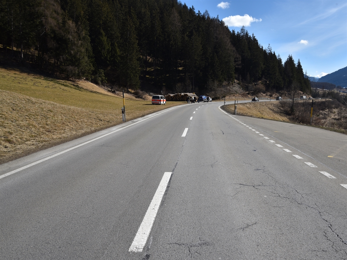 Das gerade Teilstück der Nationalstrasse N29 kurz vor einer leichten Rechtskurve. Links neben der Strasse stehen ein Patrouillenfahrzeug der Polizei sowie das beteiligte Auto und das Motorrad. Der blaue Himmel rundet das Landschaftsbild ab.