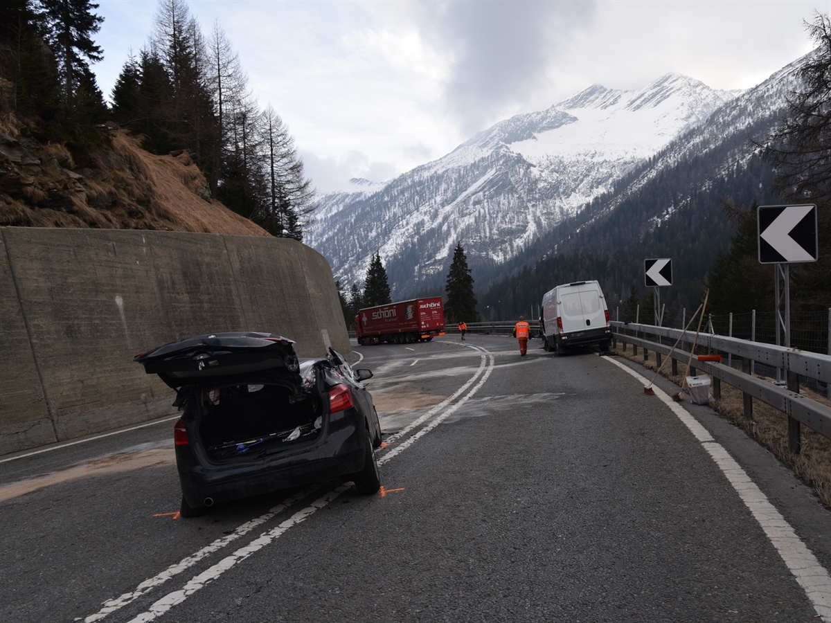 Links das total beschädigte Auto. Rechts davon im Hintergrund das Sattelmotorfahrzeug und der Lieferwagen. Hintergrund: Berge und Horizont.