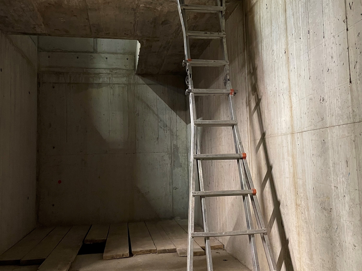 Innenaufnahme des Rohbaus. Rechts eine am oberen Betonboden angelehnte Leiter. Links oben die Aussparung des oberen Betonbodens. Unten der Bretterboden.
