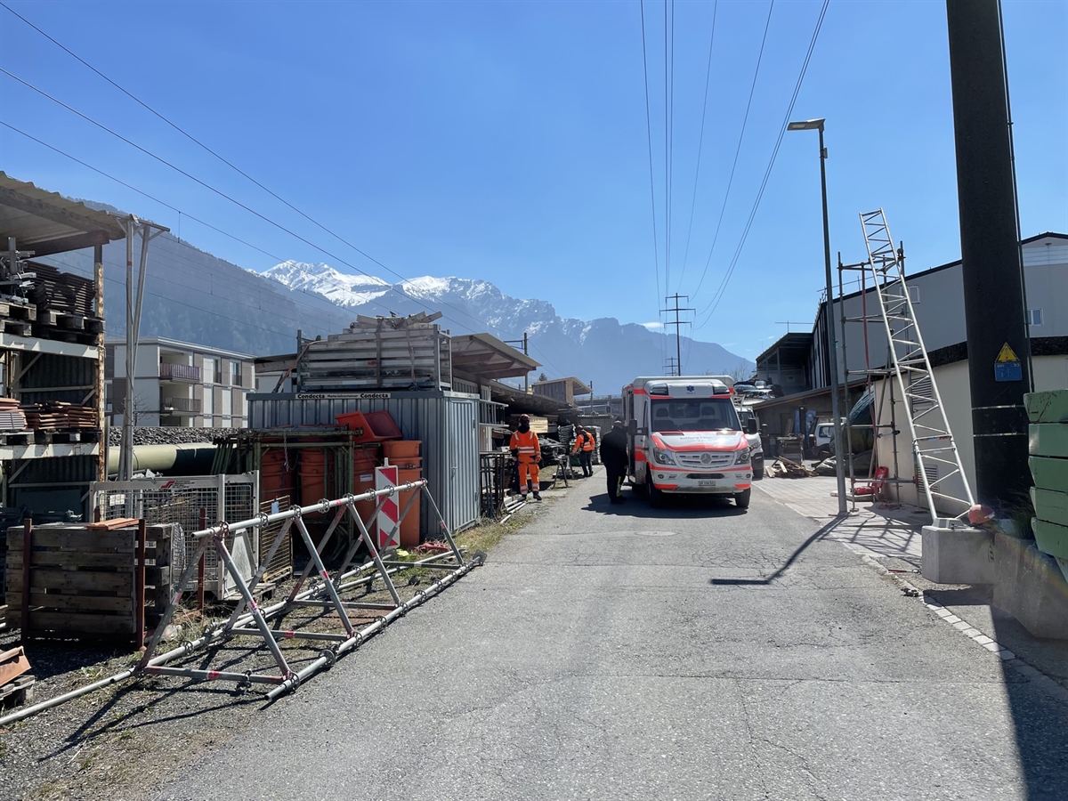 Die Unfallstelle in Zizers mit strahlend blauem Himmel. Auf der Strasse steh der Rettungswagen. Daneben befinden sich Arbeiter. Auf der linken Seite liegt der umgefallene Gerüstturm. Auf der rechten Seite steht der andere Turm, an welchem die Traverse angelehnt ist.