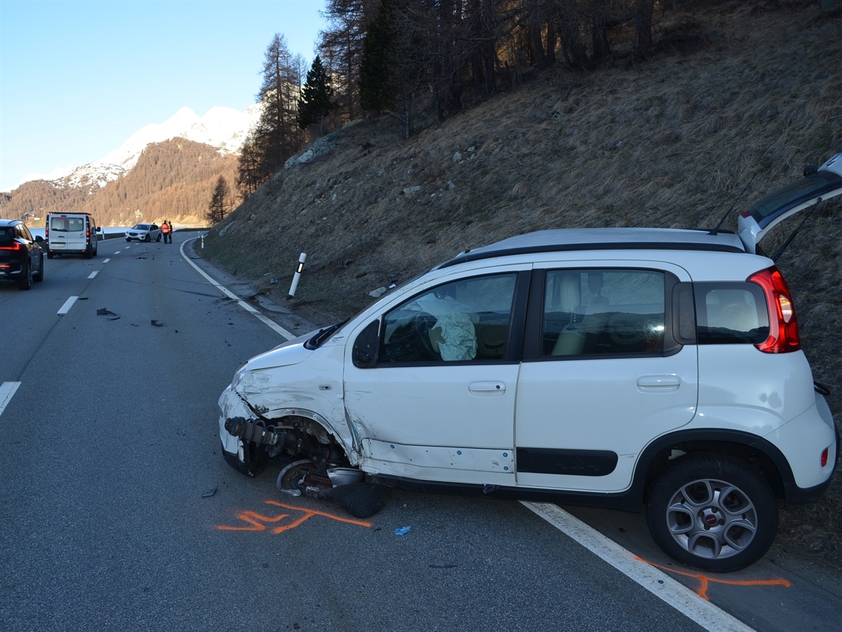 Ein weisser vorne links und seitlich beschädigter Kleinwagen steht rechts am Strassenrand. Im Hintergrund das zweite Unfallauto und Berge, welche den blauen Horizont abgrenzen.