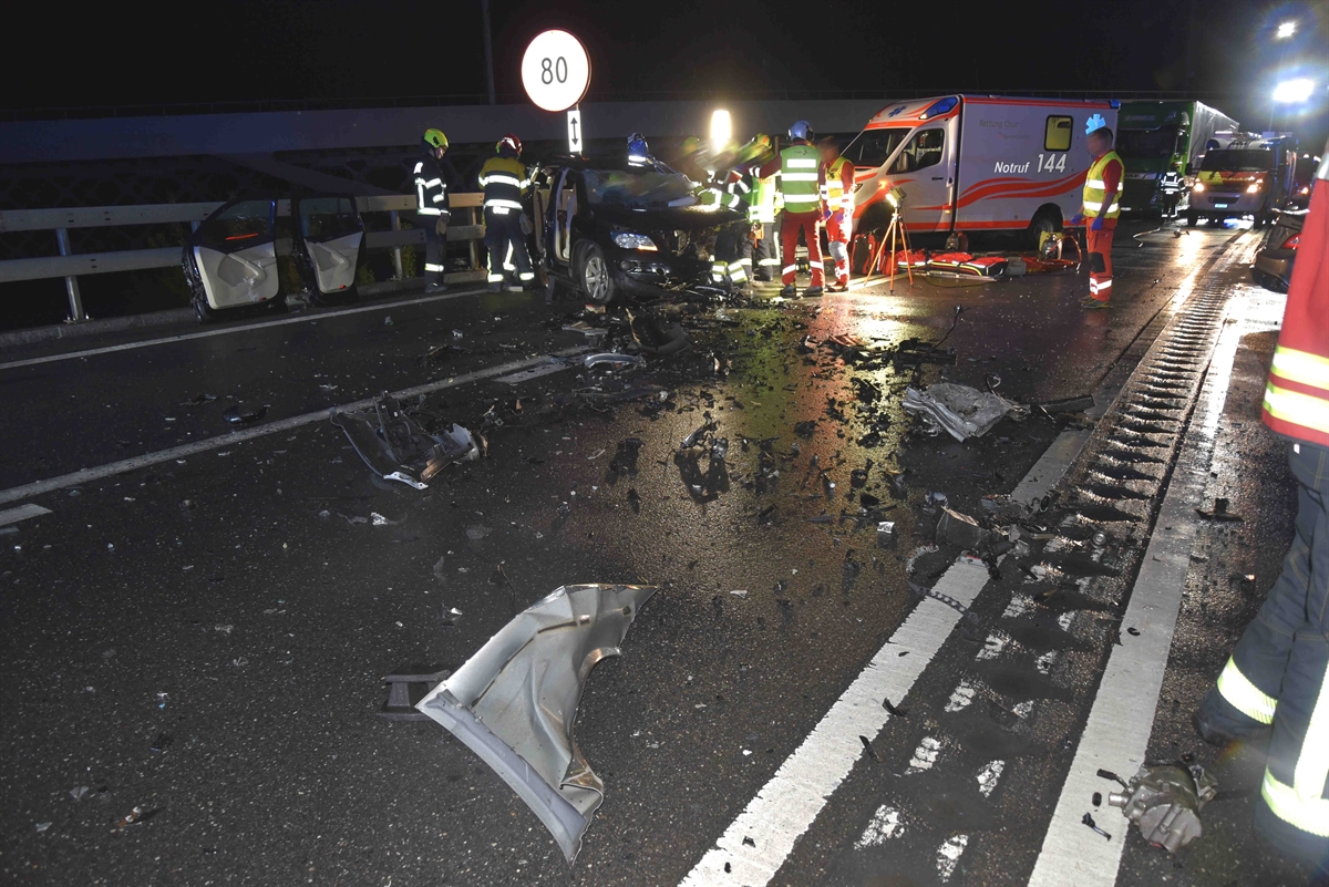 Es ist dunkel und Strasse ist nass. Diverse Fahrzeugteile liegen auf der A13 verteilt und ein total beschädigtes Unfallauto ist zu sehen. Rettungskräfte arbeiten beim Unfallfahrzeug und eine Ambulanz steht im Hintergrund.