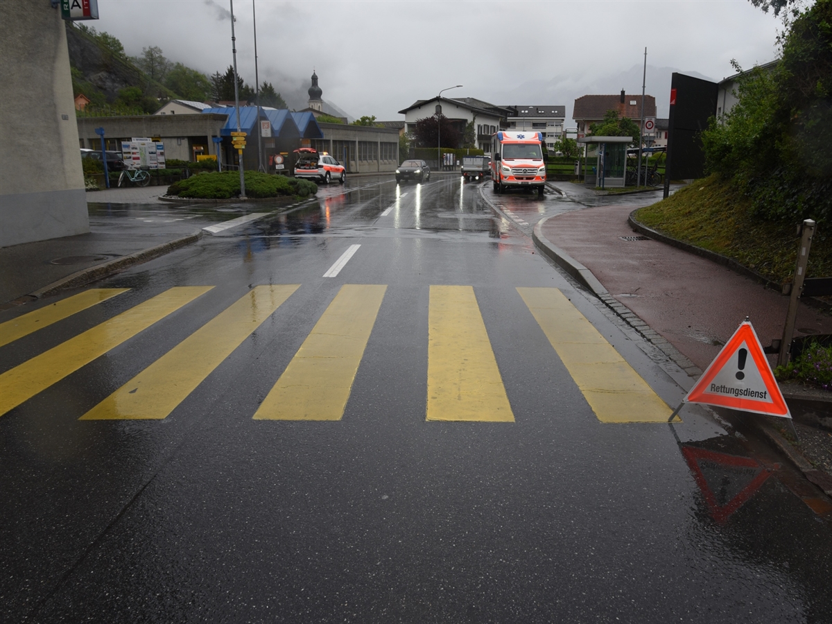 Die Hauptstrasse. Links und rechts münden Nebenstrassen ein. Rechts ein Rettungswagen, links ein Polizeiauto und im Vordergrund ein Faltsignal.