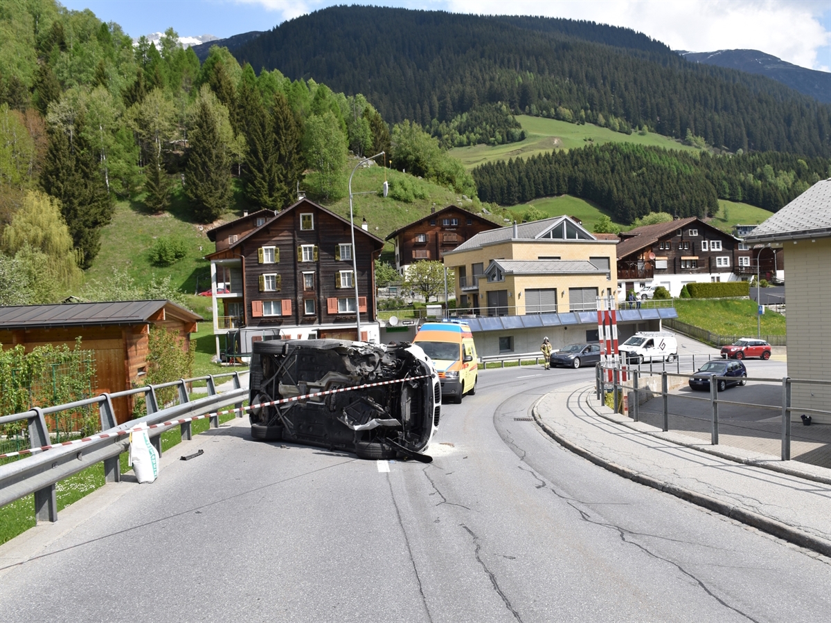 Das umgekippte Auto aus der Gegenrichtung. Hinter dem Unfallauto steht ein Rettungswagen in der Kurve. Hinter der Ambulanz hält ein Feuerwehrmann den Verkehr auf.