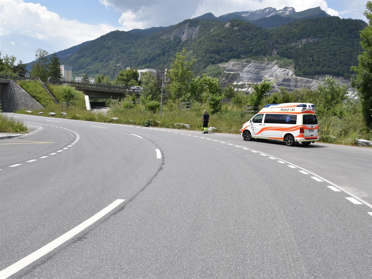Eine nach links führende Kurve, rechts auf einem abgetrennten Asphaltstreifen ein Rettungswagen. Im Hintergrund die Calanda-Berggruppe.