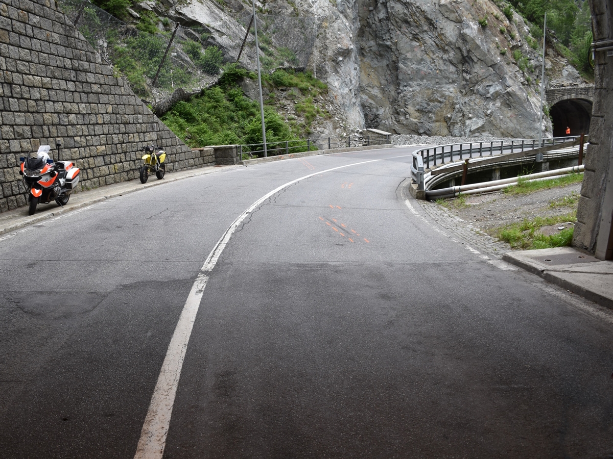 Der Tunnelausgang und die Rechtskurve. Weiter unten anderes Tunnel. Sturzspuren auf der Strasse, links vor einer Stützmauer das Unfallmotorrad und ein Polizeimotorrad.