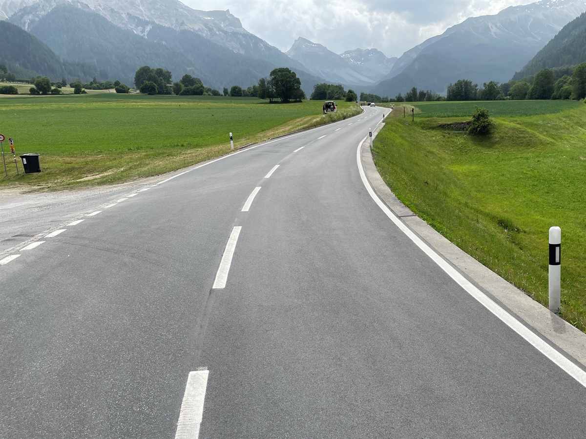 Eine Rechtskurve. Links und rechts Wiesen. Im Hintergrund Berge und der Horizont.