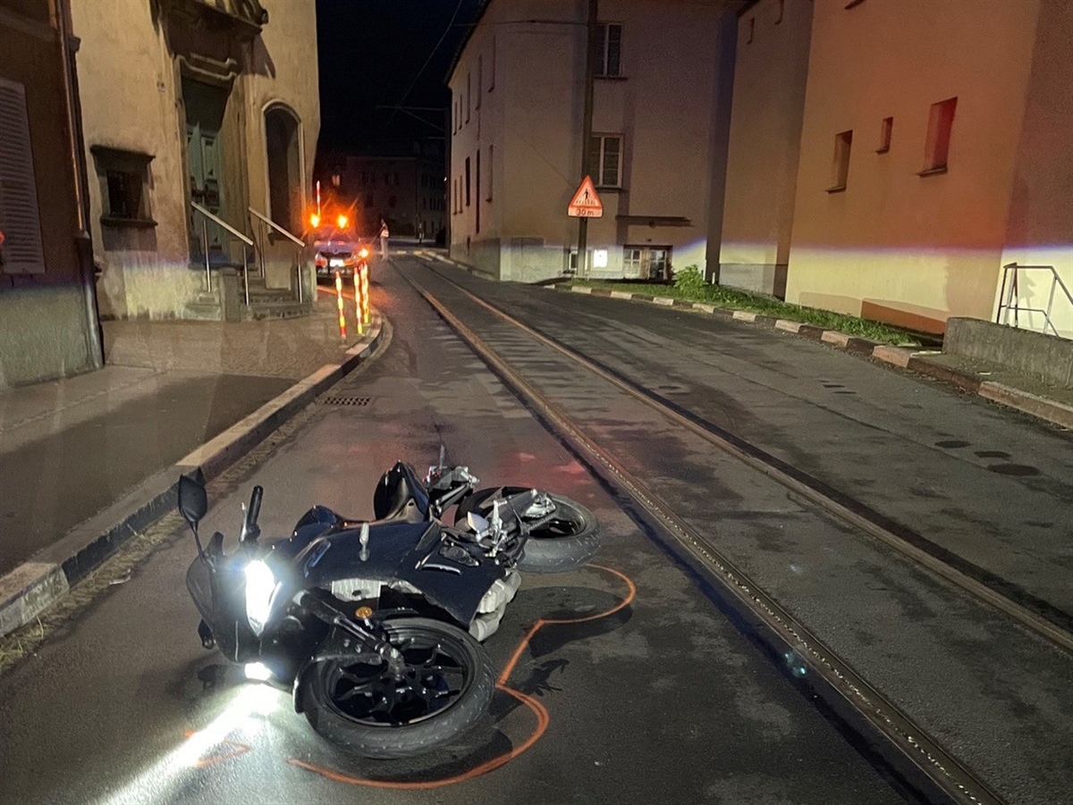 Aufnahme in der Dunkelheit. Im Vordergrund ein am Boden liegendes Motorrad. Rechts davon die Gleise. Links und rechts der Strasse Häuser was einen Engpass ergibt.